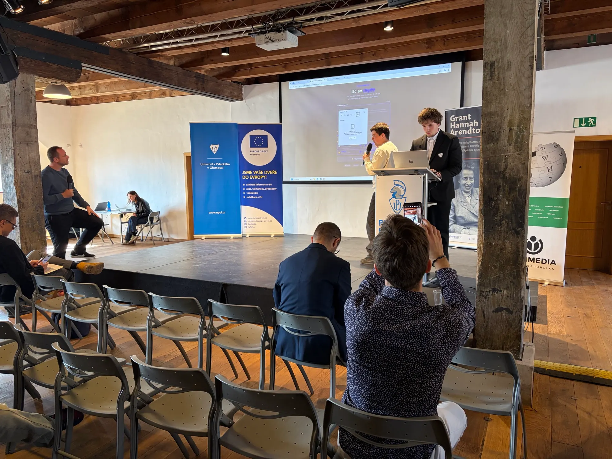 Two presenters speaking beside the competition banners